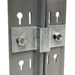 Close-up of a metal bracket connecting two vertical beams with bolts. The beams have multiple keyhole-shaped slots. The bracket is secured with two hexagonal bolts, and a cable runs through the middle between the beams.