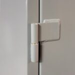 Close-up of a white door hinge attached to a light gray surface. The hinge has a simple cylindrical design with a vertical pin connection. The surrounding area is smooth and unadorned.