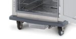Close-up of the bottom section of a stainless steel oven with an open door and metal racks inside. Beneath, a clear plastic container rests on a gray base with the Metro C5-BUMPDRIP for C5 8 and 6 Series Cabinets. Wheels are visible on the right side.