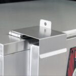 A close-up of the Metro MBQ-TRVL Travel Latch for MBQ banquet cabinets on a stainless steel box shows the edge-mounted hinge lock with a securing hole, reflecting light to highlight its sleek design.