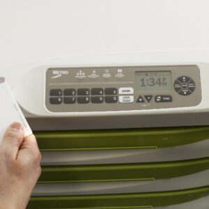 A hand holds a key card near the Metro FLX430LCDPHF Flexline High Frequency Proximity Card Reader, displaying 1:34 PM. The sleek white-and-green unit offers lock/unlock options and advanced LCD touchpad convenience.