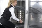 A blonde woman in a white shirt and black vest is efficiently stacking metal trays onto an industrial shelf using a Metro MBQ-SUSA Swing-Up Shelf Accessory for MBQ Banquet Cabinets.