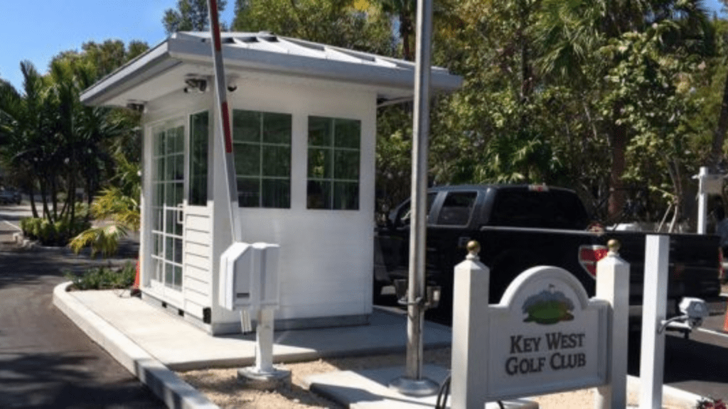 A small white guard shack with a red and white barrier arm lowered stands at the entrance. A black pickup truck waits behind. A sign in the foreground reads Key West Golf Club, nestled amidst lush greenery, exemplifying top-tier security booth customization in Guard Shack on GSA Contract