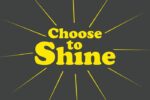 The Justrite Mat product, featuring Choose to Shine in bold yellow text on a dark gray background, displays yellow rays creating a sunburst effect that embodies positivity and inspiration.