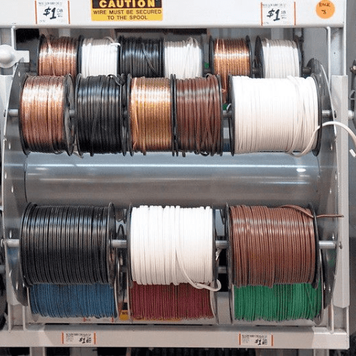 Multiple spools of electrical wire in various colors, like black, white, and red, are displayed on metal racks. Each spool is labeled with a price and a user-friendly caution sign prioritizing safety features about securing the wire is visible above.