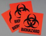 Three orange square stickers from the Justrite Label Biohazard Kit feature black biohazard symbols and the word BIOHAZARD, overlapping on a gray background, reminiscent of those found in a classic Biohazard Kit.