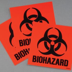 Three orange square stickers from the Justrite Label Biohazard Kit feature black biohazard symbols and the word BIOHAZARD, overlapping on a gray background, reminiscent of those found in a classic Biohazard Kit.