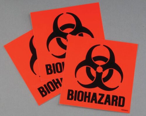 Three orange square stickers from the Justrite Label Biohazard Kit feature black biohazard symbols and the word BIOHAZARD, overlapping on a gray background, reminiscent of those found in a classic Biohazard Kit.