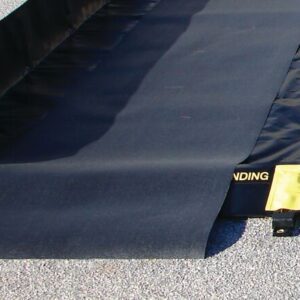 A black portable spill containment berm lies on gravel, with part of the 3X16 Justrite Track Mat unfolded. A textured mat shows a LANDING section in yellow beside a yellow tag.
