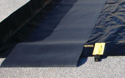 A black portable spill containment berm lies on gravel, with part of the 3X16 Justrite Track Mat unfolded. A textured mat shows a LANDING section in yellow beside a yellow tag.