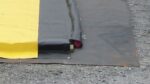A 6X28 Justrite Ground Mat is unfurled on gravel, displaying a red section. Metal grommets edge the yellow and black tarp, boosting its durability and practicality.