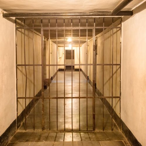 A dimly lit, narrow hallway with ballistic glass for guard shacks in the foreground. The corridor has a concrete floor and white walls, leading to a single closed door at the end. The space exudes an austere and confined appearance.