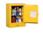 The yellow Justrite Flam Mini 2G Cabinet stands with its door slightly ajar, revealing shelves stocked with spray cans and bottles. Hazard warning symbols adorn the front, while a safety information label is affixed inside.