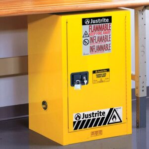 The Justrite Cabinet, Flam Cmp 12G, Sc Yel is placed under a table and features bold warnings like FLAMMABLE KEEP FIRE AWAY, INFLAMMABLE, along with a black and white hazard symbol, providing safety with its lock feature. Ideal for storing flammable liquids.