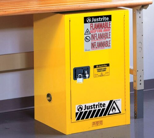 The Justrite Cabinet, Flam Cmp 12G, Sc Yel is placed under a table and features bold warnings like FLAMMABLE KEEP FIRE AWAY, INFLAMMABLE, along with a black and white hazard symbol, providing safety with its lock feature. Ideal for storing flammable liquids.
