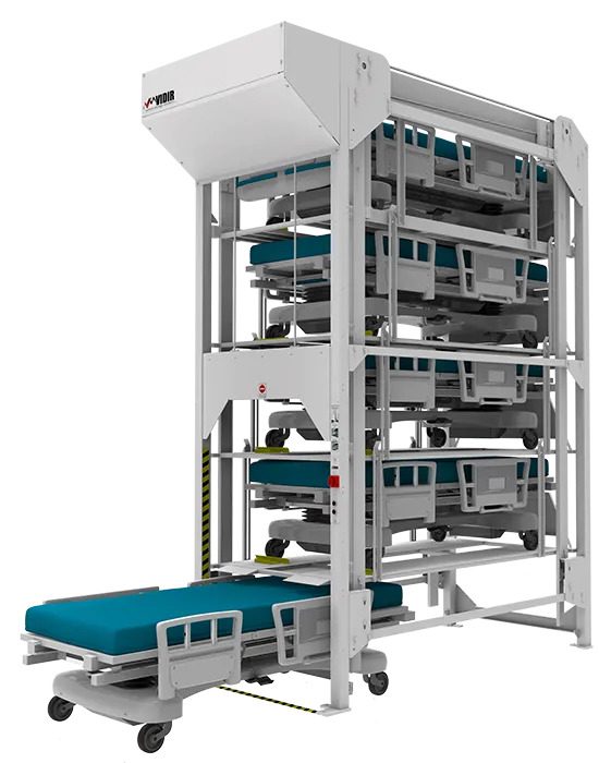 A vertical bed storage system with multiple levels, featuring blue mattresses on hospital beds stacked within a white metal frame. The Vidir Hospital Bedlifts platform extends from the lower level, showcasing space-saving hospital bed storage solutions.