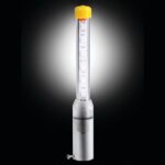 The Justrite Whip features a transparent cylindrical LED light in a clear tube with a yellow top, connected to a metallic base via a small screw. Its vivid glow contrasts sharply against the black backdrop.