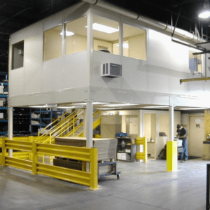 An industrial warehouse space houses a large, elevated white office structure known for fast construction modular offices. It features windows and a yellow staircase leading up to it. A person works nearby, with safety barriers ensuring security around the site.