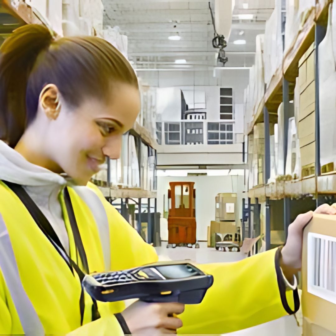 A woman in a yellow safety vest smiling while scanning a barcode with a handheld device in a warehouse setting