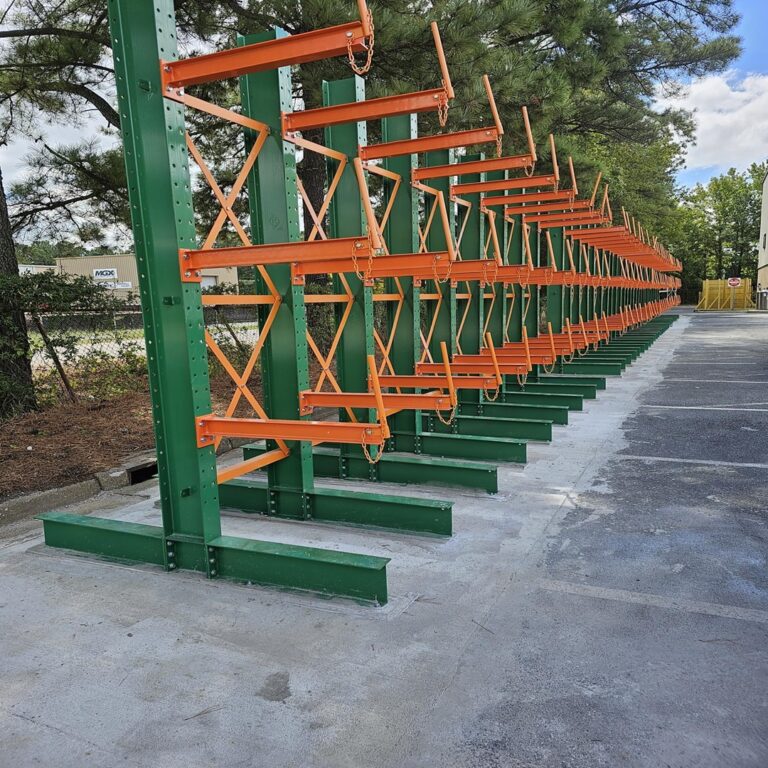 A row of green and orange selective pallet racks extends along a concrete path outdoors. Trees and a fence border the area, with a cloudy sky in the background. These racks are likely used for industrial storage.
