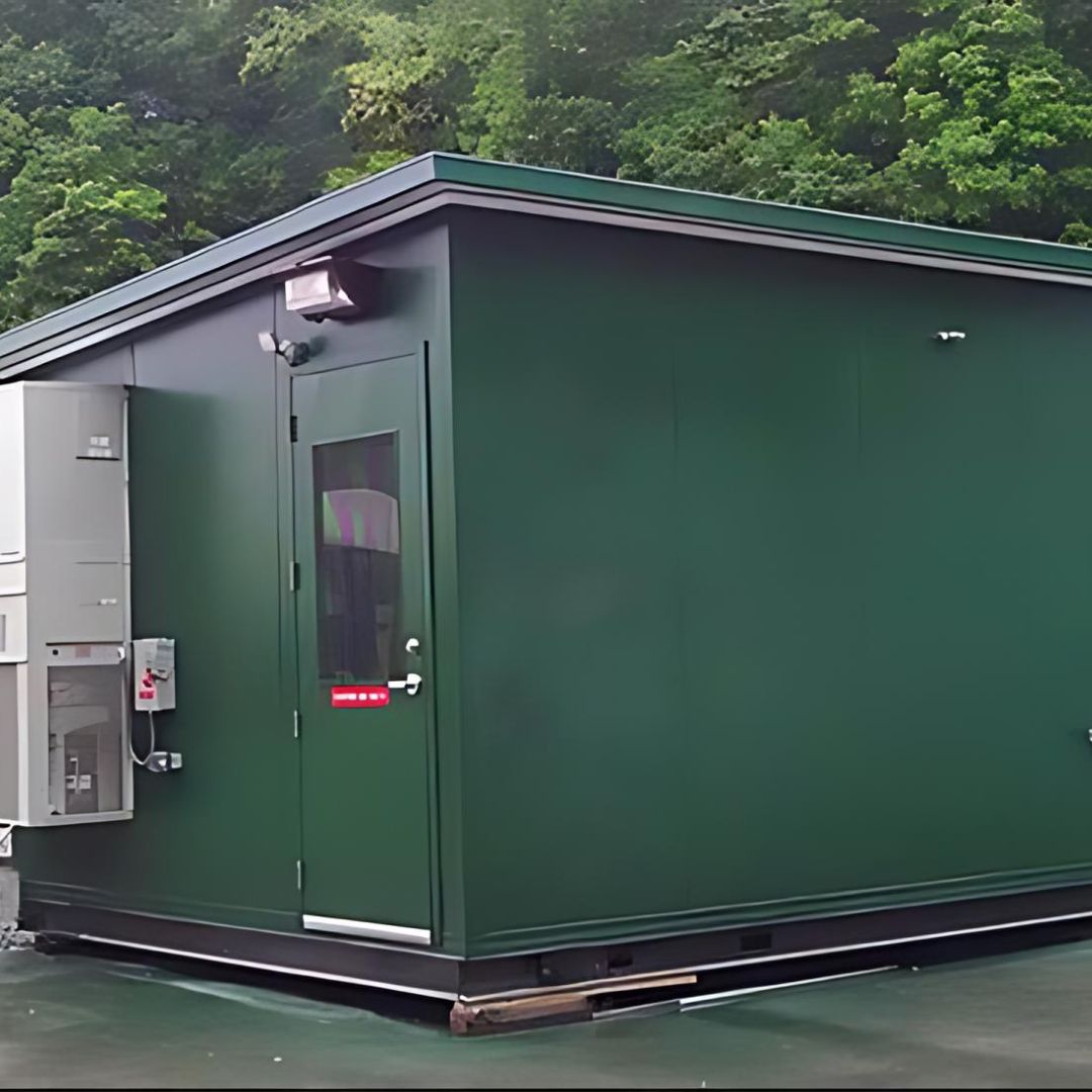A dark green outdoor machine enclosure with HVAC and electronic panels mounted to one side. It features a single door with a window, security lighting above, and is situated on a temporary base.