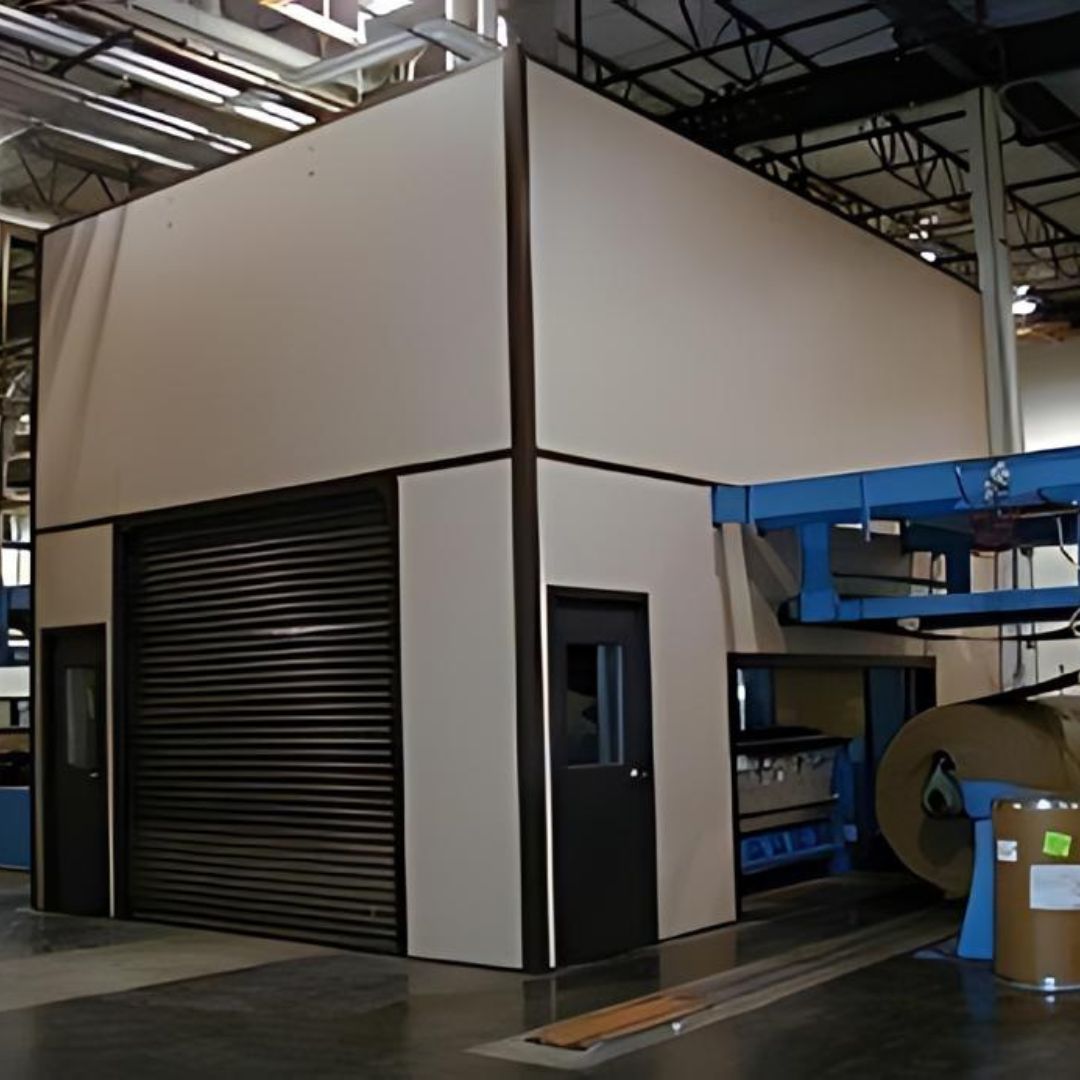 A large, beige machine enclosure installed indoors, with a black roll-up door and a standard door for access. It has contrasting dark trim and is positioned next to industrial equipment, suggesting its use in a manufacturing setting.