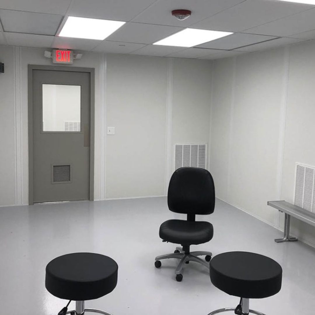 Inside a white modular cleanroom with a vision-panel door, stools and a chair