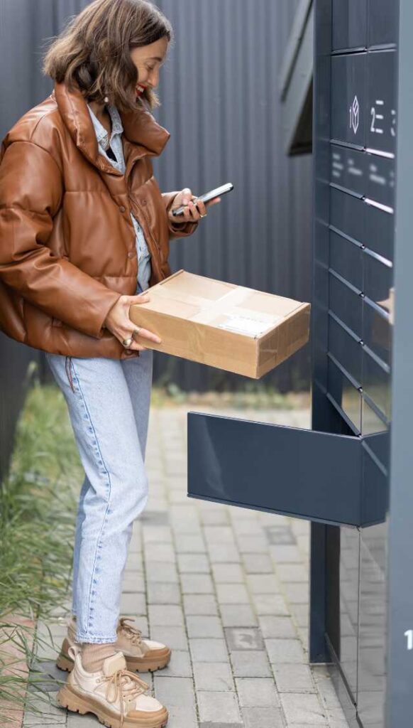 Lady wearing a brown jacket and white pants holding her mobile phone on the left and a parcel on the right , standing in front of a parcel locker.
