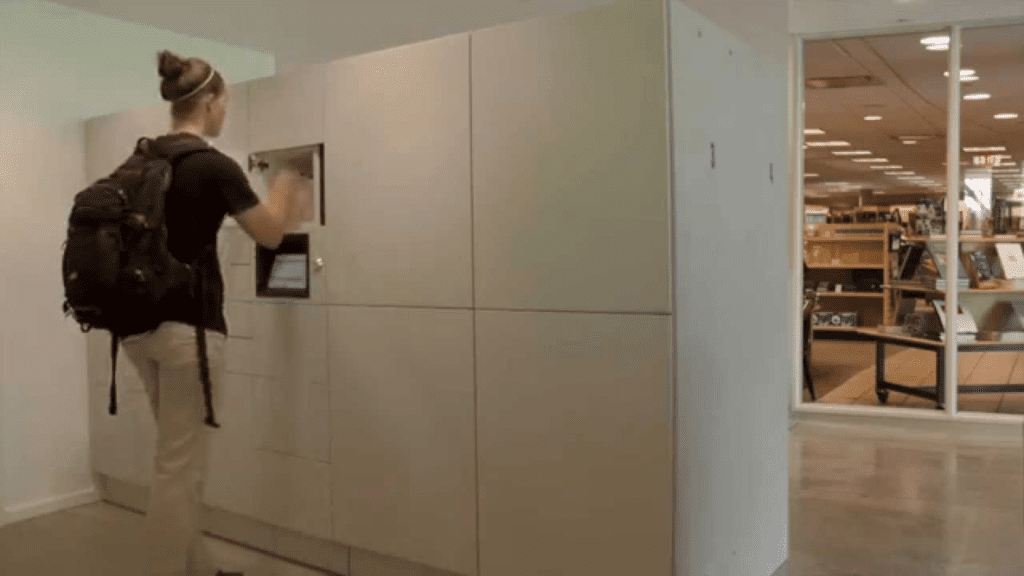 A person with a backpack interacts with sleek, secure parcel lockers in a modern interior. A glass wall unveils a well-lit room with bookshelves and a table, evoking a university library atmosphere.