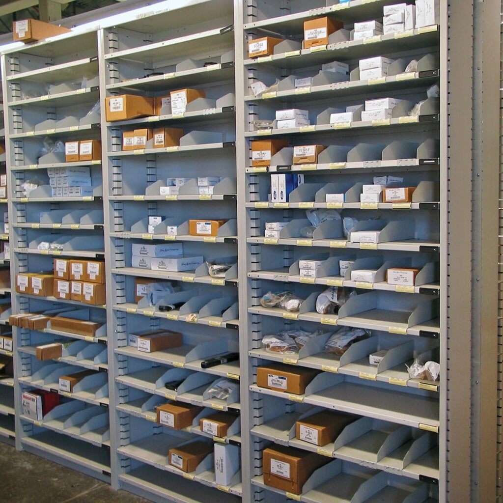 A storage area with tall metal shelves features bin shelving, where various small packages, boxes, and labeled containers are organized in compartments. The shelves are mostly full, with labels aiding in identification and organization.