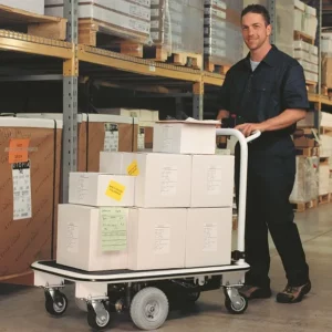 In a warehouse aisle, someone in a dark uniform smiles behind an Electro Kinetic Technologies Pony Express Motorized Platform Truck, 1500 lb. capacity, with large white boxes on its 40 L x 25 1/2 W deck. Shelves filled with more boxes highlight the trucks efficiency.