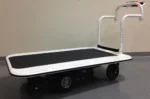 A white flatbed cart with black wheels and a curved handle features a black non-slip platform. With a 3000 lb. capacity, it stands on a gray speckled floor by a light wall, set to tackle tasks like the Electro Kinetic Technologies Pony Express Motorized Platform Truck.