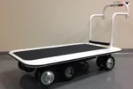 A Pony Express Motorized Platform Truck by Electro Kinetic Technologies features a black surface, six wheels, and a white handlebar on a gray floor against a beige wall. With its 52 L x 34 1/2 W deck and robust 4000 lb. capacity, its ideal for heavy-duty tasks.