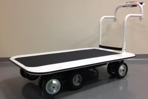 A Pony Express Motorized Platform Truck by Electro Kinetic Technologies features a black surface, six wheels, and a white handlebar on a gray floor against a beige wall. With its 52 L x 34 1/2 W deck and robust 4000 lb. capacity, its ideal for heavy-duty tasks.