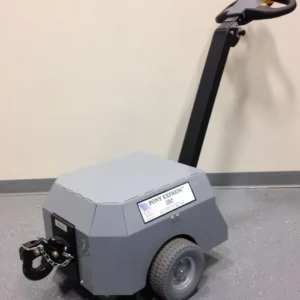 A small, gray motorized cart with a handle and two large wheels rests on the floor labeled Electro Kinetic Technologies, Pony Express Electric Tugger. Equipped with a Pintle & Hook Hitch, it offers a robust 5000-pound capacity for heavy-duty tasks.