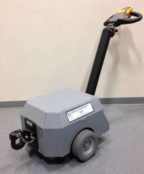 9894db16-1c62-434a-8fb5-61249a76f78d_zpsac786792.webp A small, gray motorized cart with a handle and two large wheels rests on the floor labeled Electro Kinetic Technologies, Pony Express Electric Tugger. Equipped with a Pintle & Hook Hitch, it offers a robust 5000-pound capacity for heavy-duty tasks.