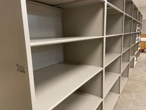 Empty beige metal shelving units, reminiscent of those at the Utah Museum, are lined up against a wall in a room with a concrete floor. The shelves are clean and uniformly spaced, creating a neat and orderly appearance.