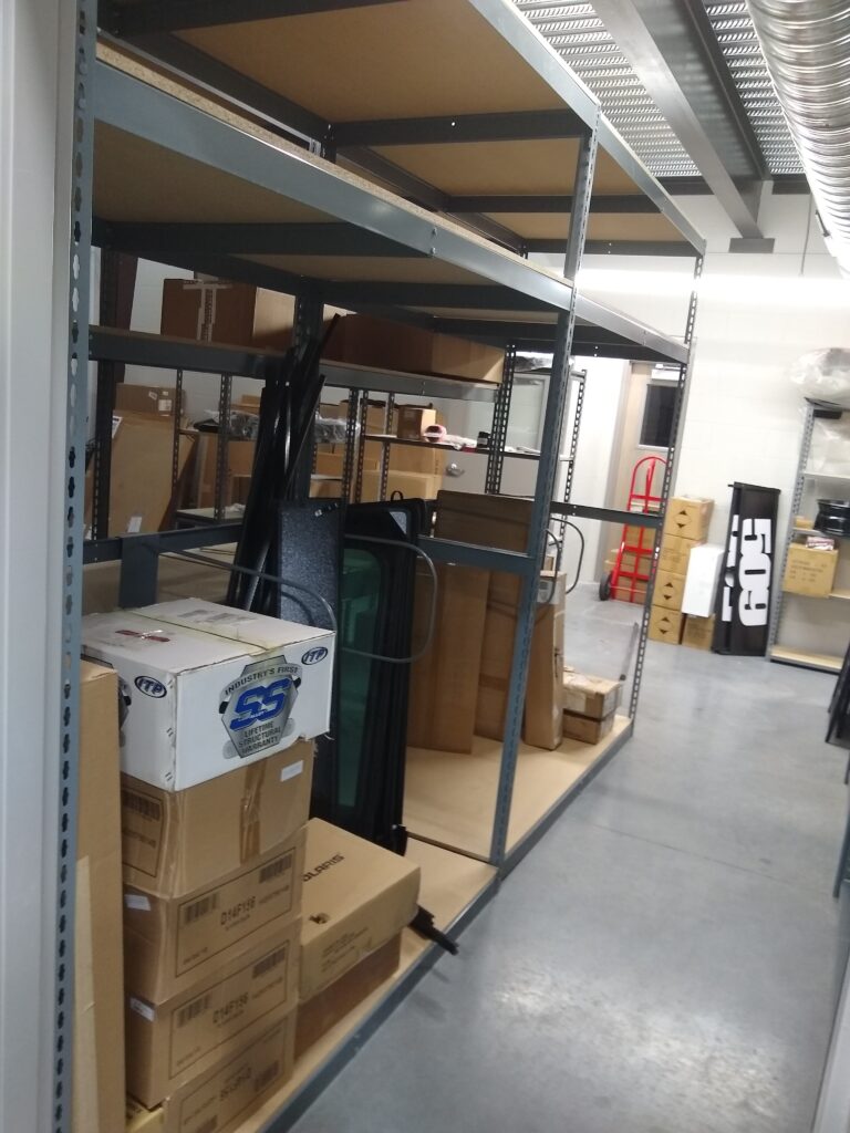 The storage room features customizable shelving solutions with metal shelves holding cardboard boxes of various sizes. Black metal pieces lean against the shelves, and a red hand truck can be seen in the background, perfect for heavy-duty parts storage. The gray floor is polished and clean.