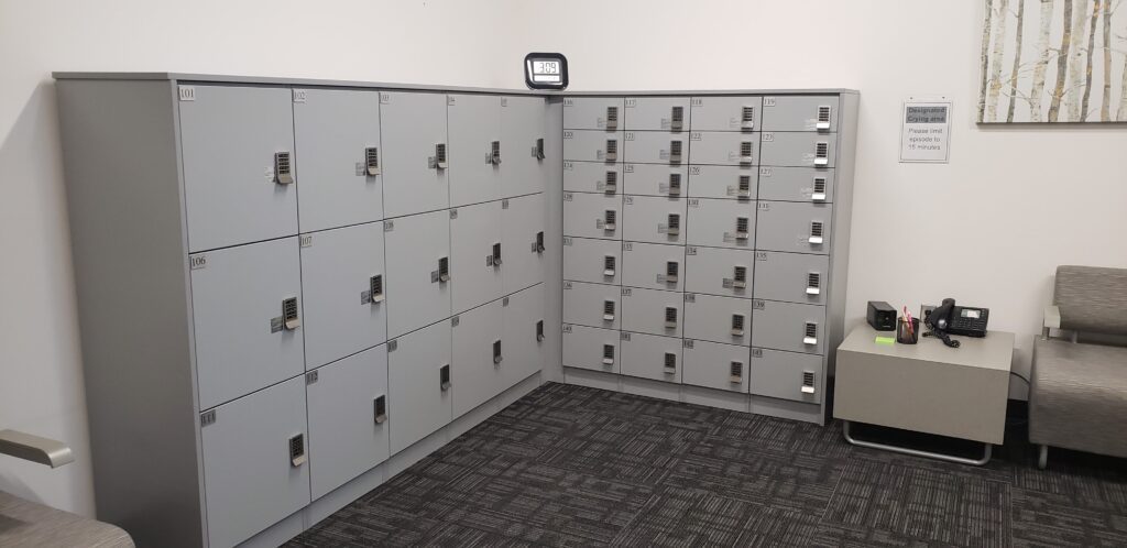 The Utah State University Testing Center features an L-shaped array of gray lockers with combination locks, supplied by Material Handling USA for secure locker storage. A digital clock rests on top, while a small table with a phone and a couch completes this custom locker design.