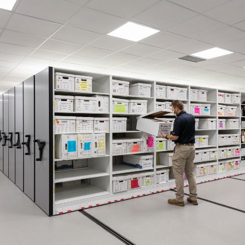 A person is organizing boxes on large, labeled storage shelves in a bright, spacious room. The shelves are filled with uniform boxes critical for forensic evidence storage solutions, and the smooth, clean floor enhances the efficiency of what appears to be an archive or storage facility.