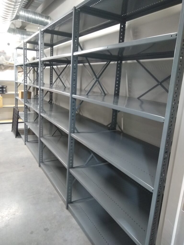 Heavy-duty parts storage shelves line the gray concrete floor of a storage room, their metallic frames standing sturdy beneath visible air ducts on the ceiling. Each shelf is evenly spaced and supported, perfect for powersports parts shelving.