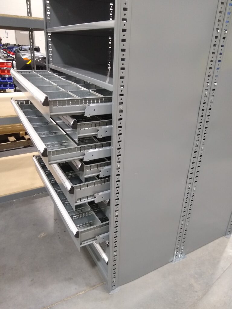 Steel shelving unit with several open metal drawers, each containing multiple compartments. Ideal for Powersports Inventory Management, the unit is in a storage room with a concrete floor, providing customizable shelving solutions to optimize space and organization.
