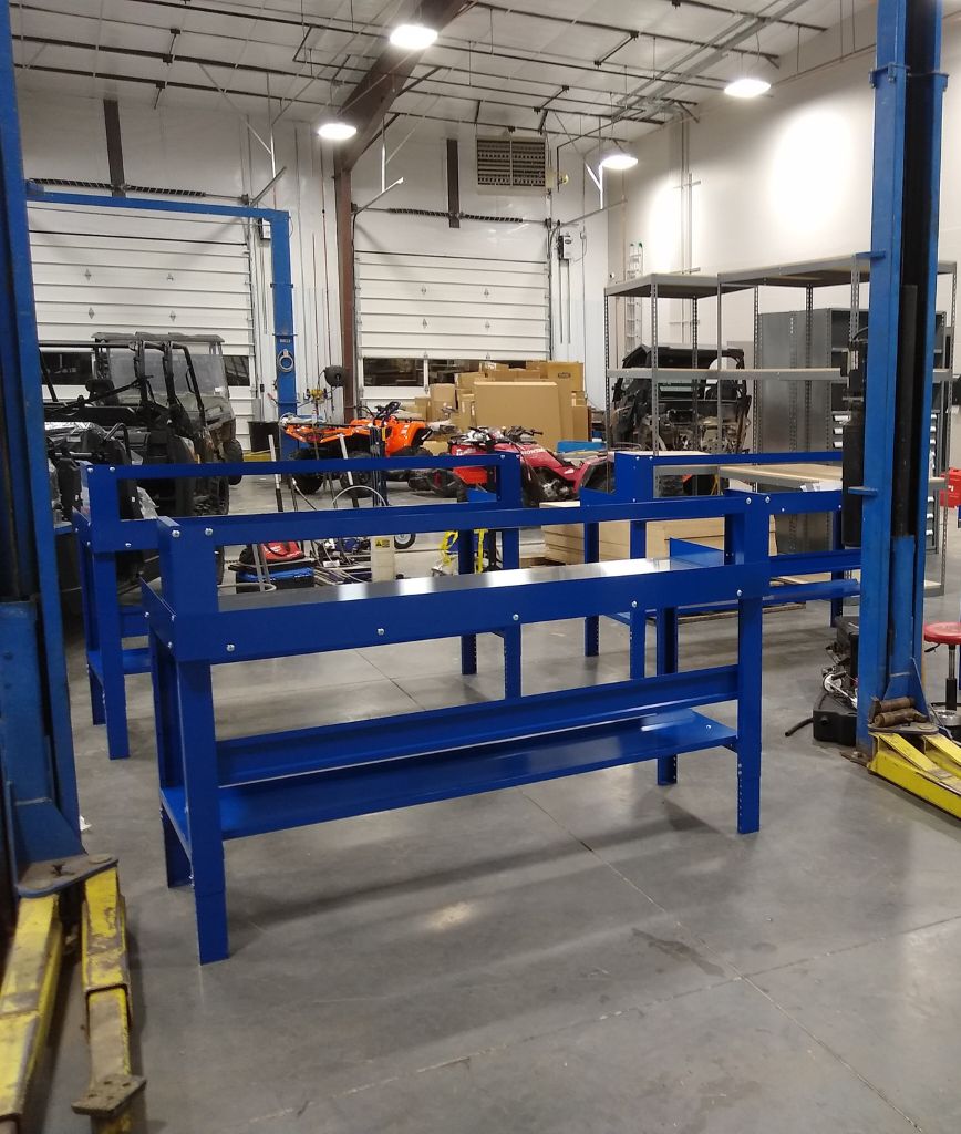 A spacious workshop with blue metal tables, customizable shelving solutions filled with various tools, and equipment surrounding vehicles in the background. The room is well-lit with fluorescent lights, and a garage door is partially open.