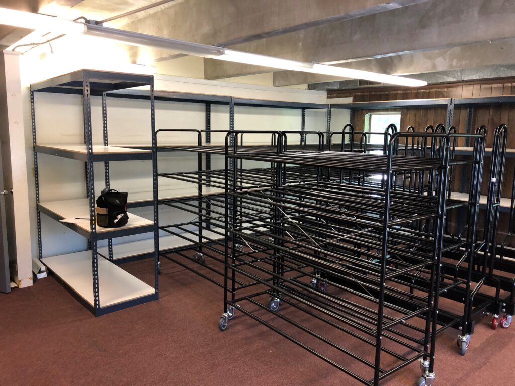 A room with several metal storage racks, some featuring mobile shelving and white shelves. Many racks are empty and stacked on top of each other. A black bag rests on one shelf. The room is accented by a brown carpet, wooden panel walls, and a concrete ceiling.