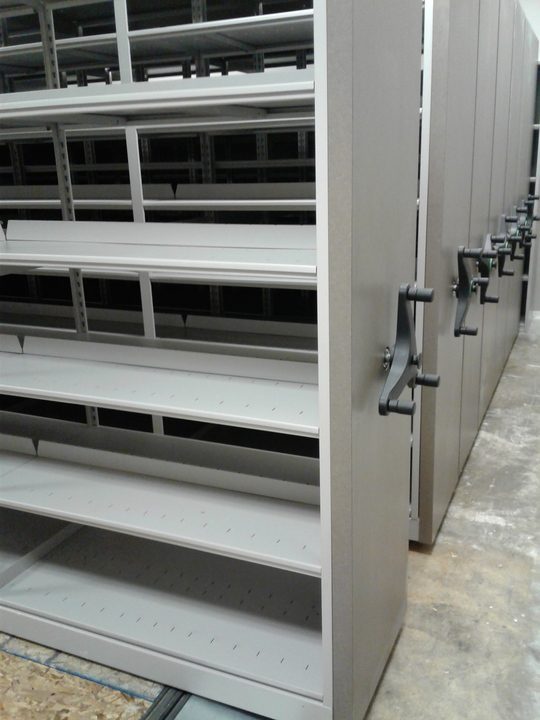 Rows of mobile shelving units with visible crank handles line the storage area. The mostly empty shelves highlight the efficient use of space, while the floor is crafted from a light-colored material, enhancing the areas brightness.