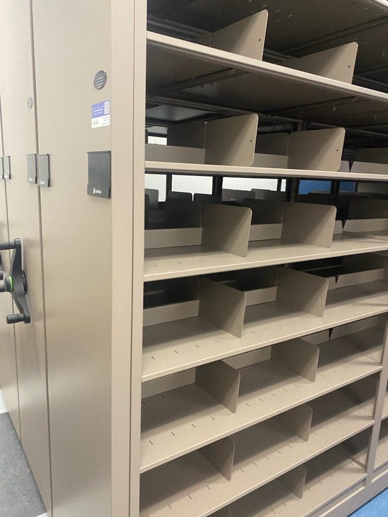 A large beige storage cabinet with open shelves and metal dividers features a wheel handle, emphasizing its versatility as a mobile shelving system. The shelves remain empty in a room with a gray floor, showcasing its potential to adapt to various organizational needs.