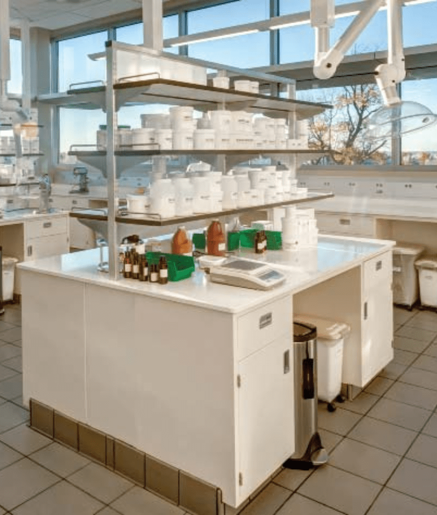 A modern laboratory with white countertops and shelves stocked with containers and bottles. There are windows in the background letting in natural light. The lab is clean and organized, with various equipment and storage bins visible.