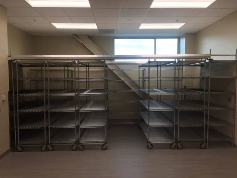 Rows of mobile high-density shelving units stand ready in a room with beige walls, linoleum floors, and fluorescent lights. A window on the back wall lets natural light stream in, highlighting their versatility and efficiency for mobile high density shelving