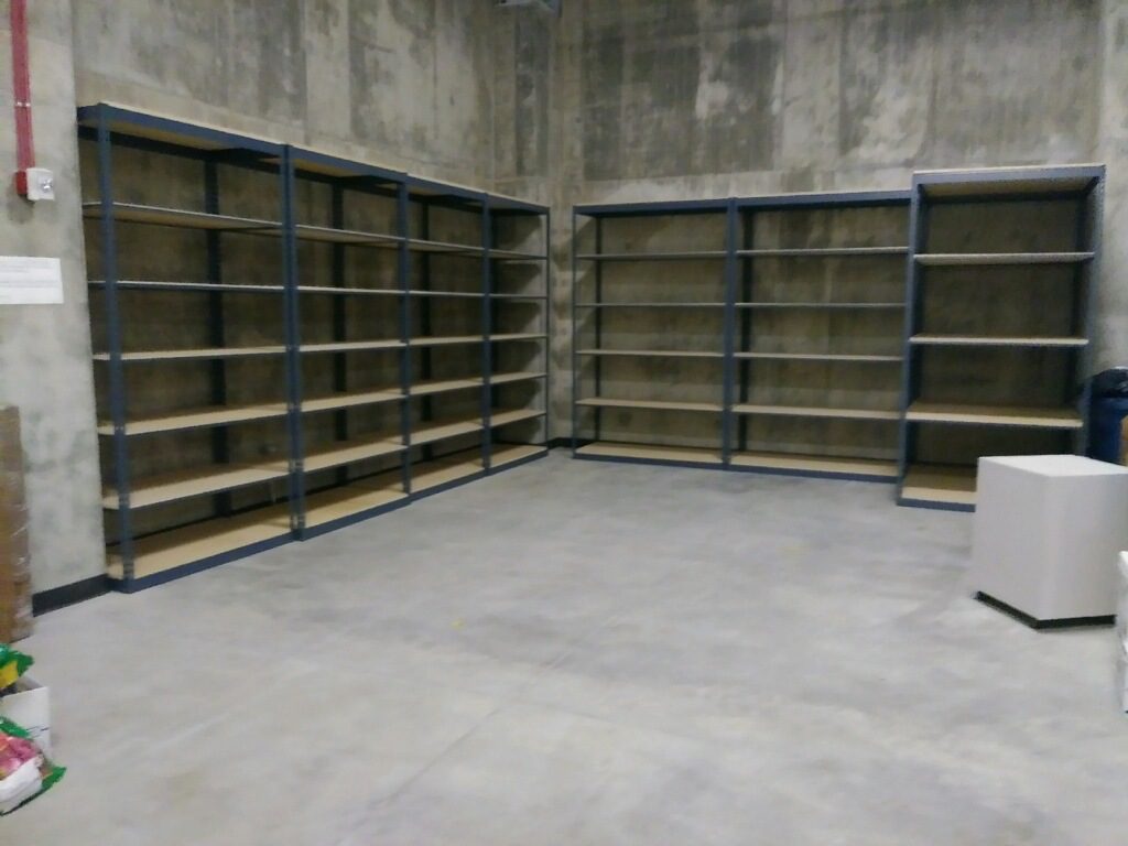 An empty storage room with concrete walls and a sturdy floor houses two sets of metal shelving units in the corner. These static shelves contrast with the potential efficiency of mobile shelving. Nearby, a small white cabinet rests on the right side, adding to the sparse decor.