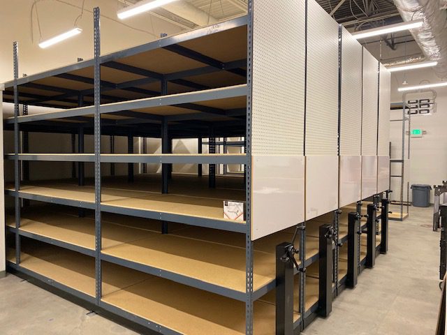 Rows of wide-span boltless shelving with white end panels and full-width particle board decks in the Tecovas stockroom